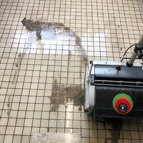 Ein Bodenreiniger mit grün-rotem Aufkleber steht auf einer Fliesenfläche, auf der Wasser und Schmutz zu sehen sind.