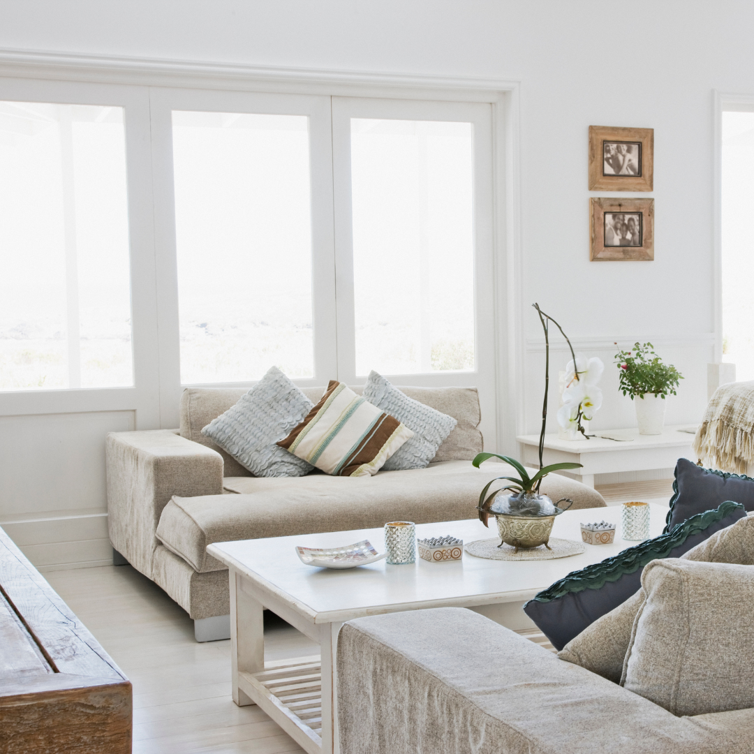 Ein helles, modernes Wohnzimmer mit einem beigen Sofa, einem weißen Couchtisch, dekorativen Kissen und Pflanzen, sowie großen Fenstern und Bilderrahmen an der Wand.