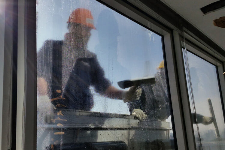 Ein Fensterreiniger in orangefarbener Mütze und Handschuhen wischt ein Fenster mit einem Schwamm ab.